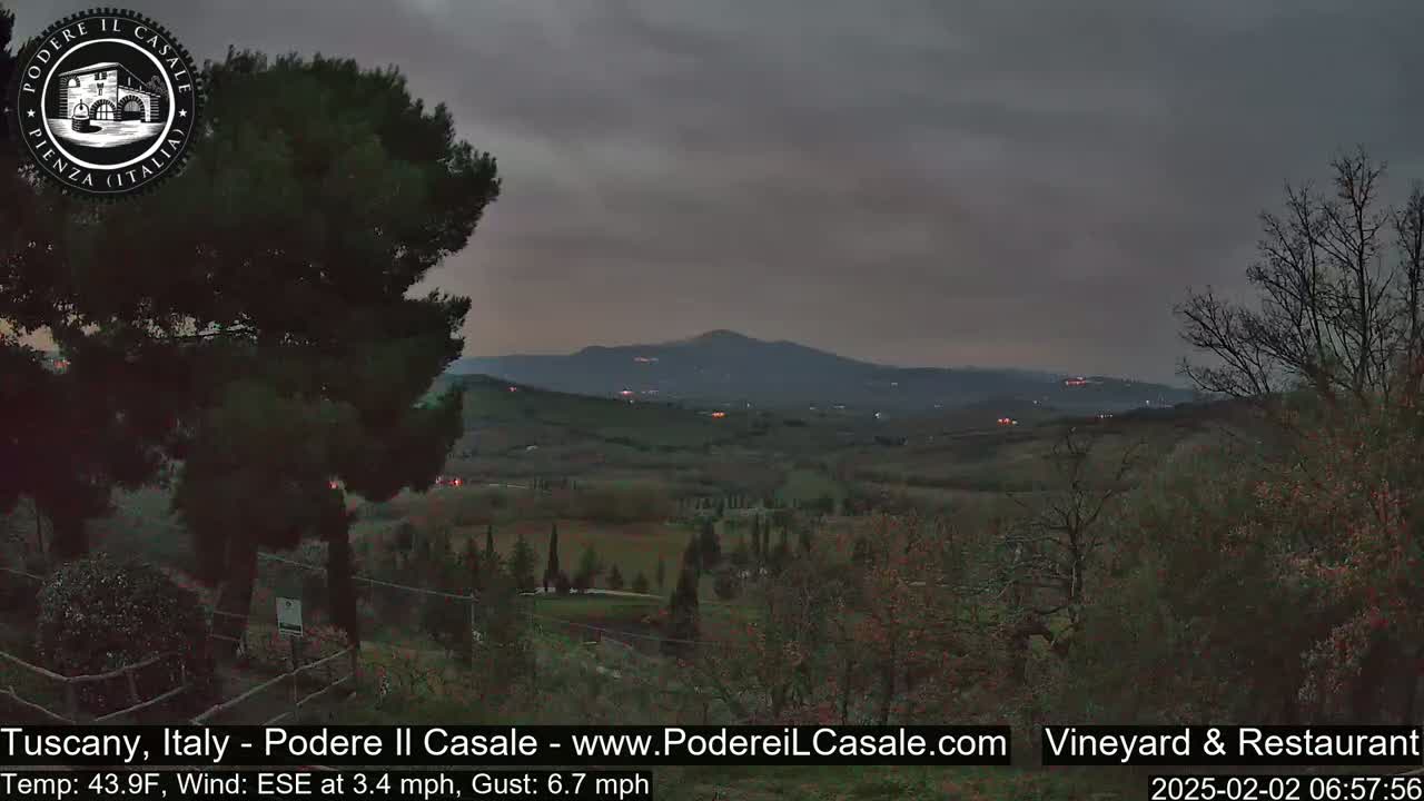 A view of a Tuscan hillside at twilight under a cloudy sky, showing rolling hills, sparse trees, and distant lights.