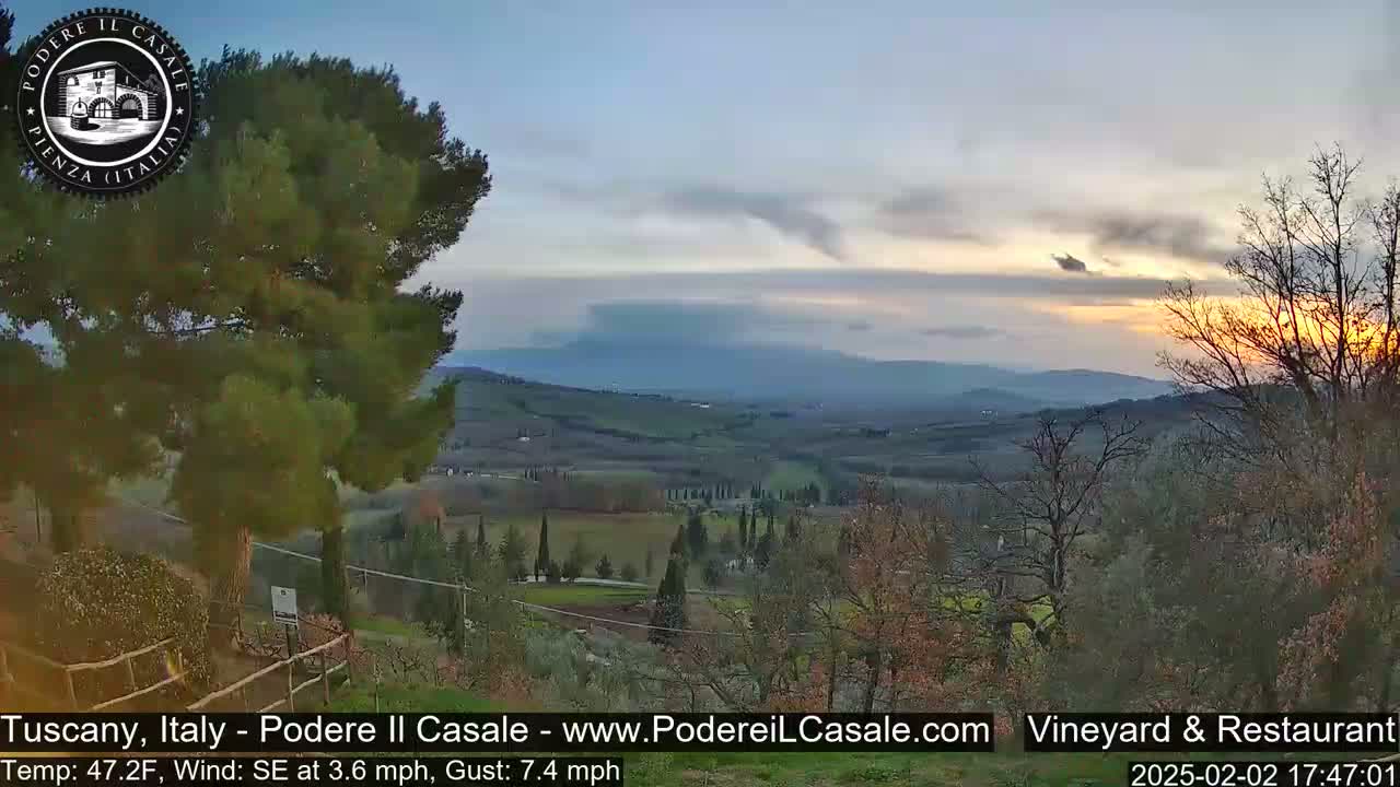 A sunset over a Tuscan landscape, showing rolling hills, vineyards, and trees under a partly cloudy sky.