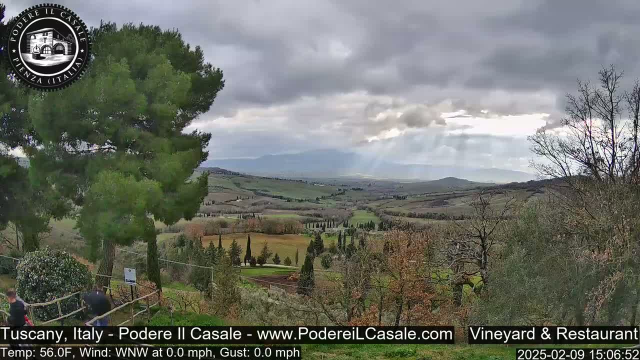 A view of a rolling Tuscan landscape under a cloudy sky, with sunbeams breaking through the clouds.