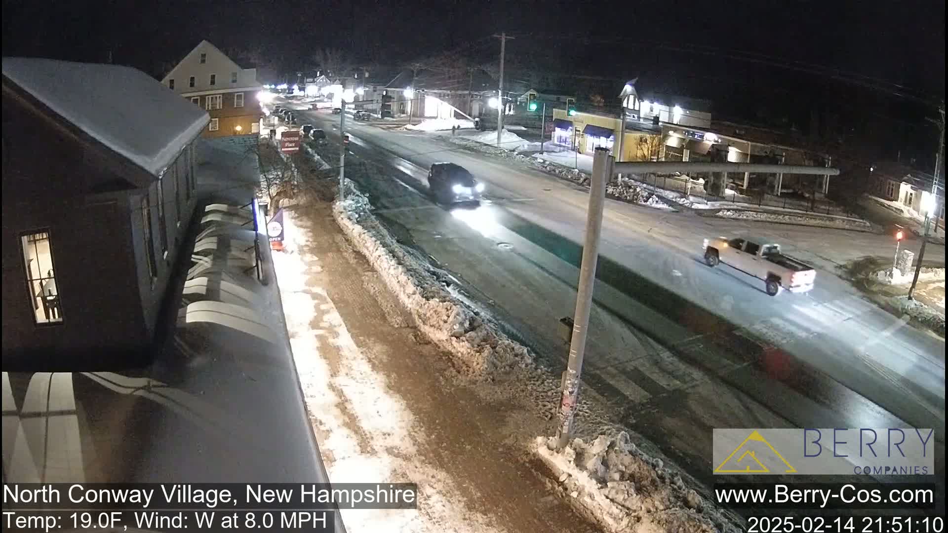 A snow-covered street at night shows several cars driving down a road lined with buildings, under a dark sky.