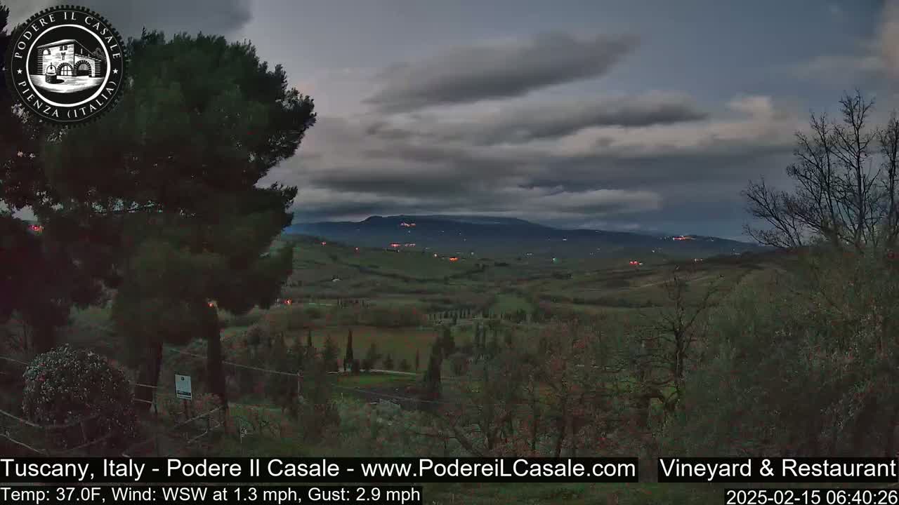 A dark, overcast sky hangs over a gently rolling Tuscan landscape dotted with sparse trees and the faint lights of distant buildings.