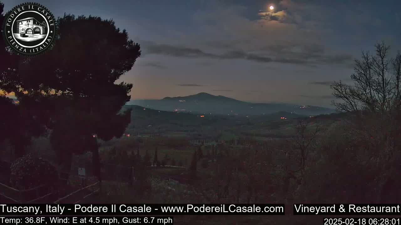 A nearly full moon shines in a twilight sky over a dark, hilly landscape dotted with sparse, low-lying vegetation and distant, faint lights.