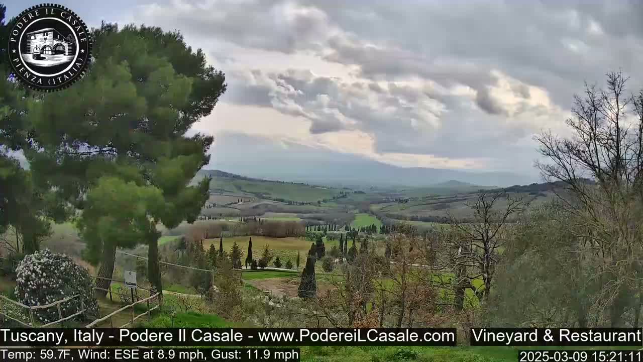 A partly cloudy Tuscan landscape with rolling green hills, dotted with trees and shrubs, is visible from an elevated vantage point.
