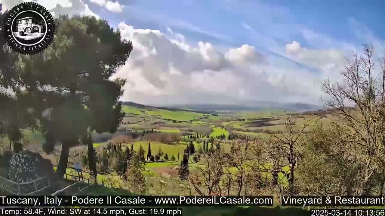 A partly cloudy, sunny day reveals a picturesque Tuscan landscape of rolling green hills, dotted with trees and farmland, viewed from a high vantage point.