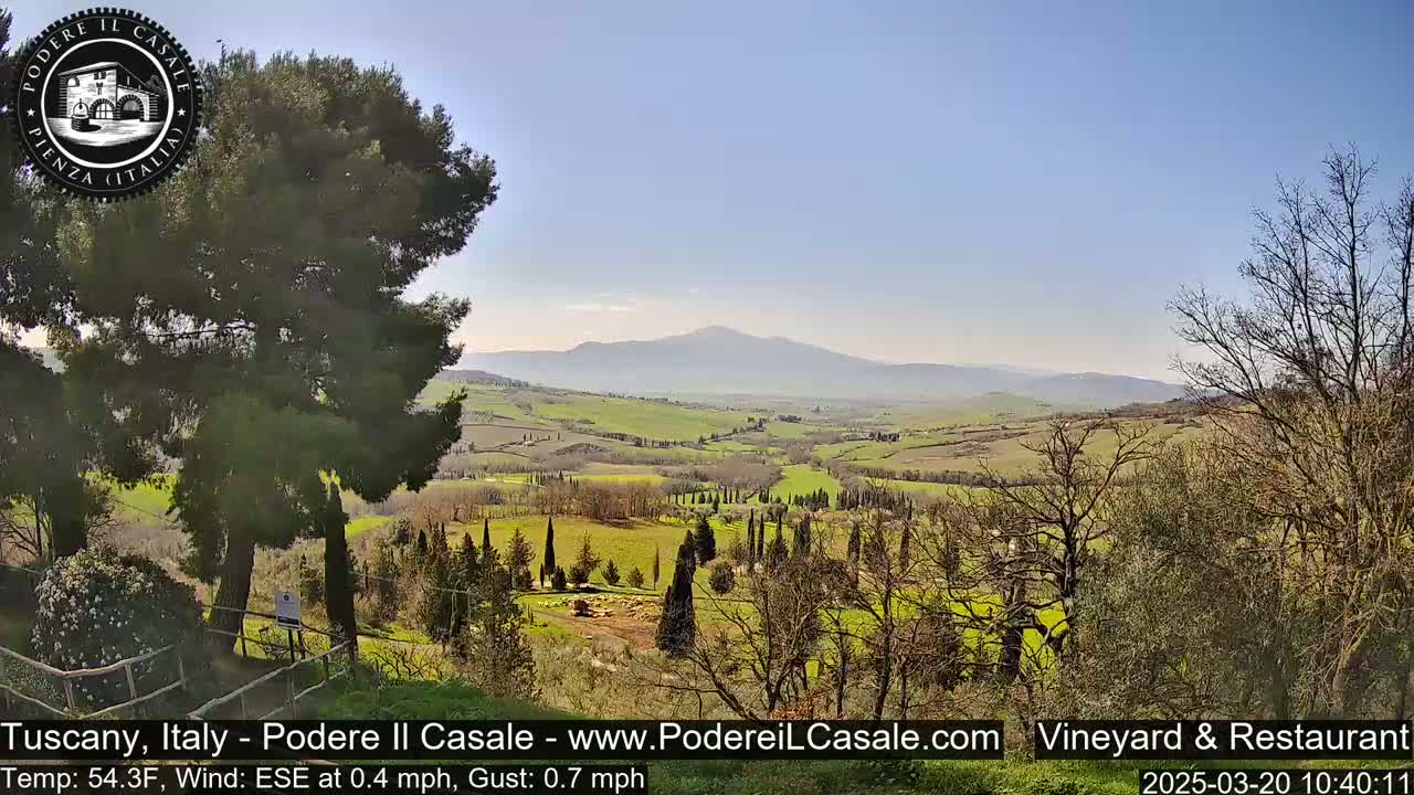 A panoramic view of a sunny Tuscan countryside reveals rolling green hills, cypress trees, and a distant mountain range under a clear blue sky.