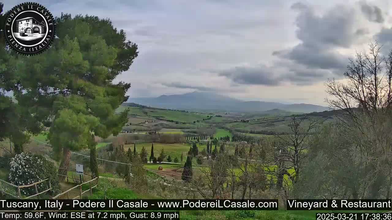 A partly cloudy Tuscan landscape shows rolling green hills dotted with trees and a distant mountain range.