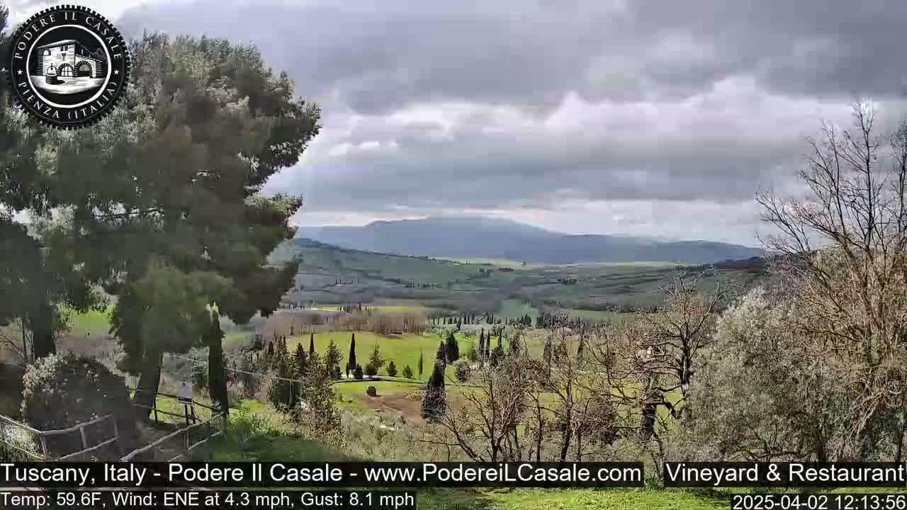 A partly cloudy sky overlooks a gently rolling, green Tuscan landscape dotted with trees and farmlands.