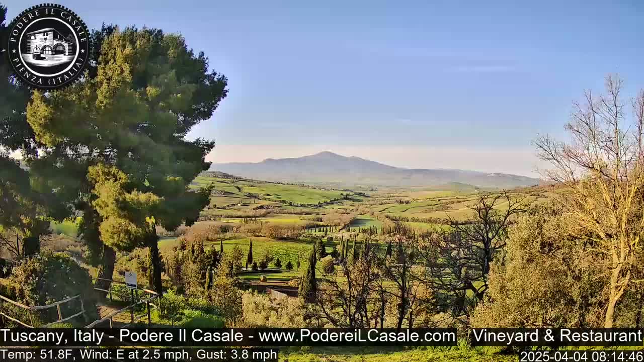 A sunny, partly clear day reveals a picturesque Tuscan landscape with rolling green hills, vineyards, and a distant mountain range.