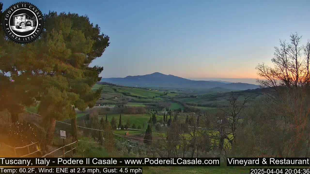 A sunlit Tuscan landscape shows rolling green hills and a distant mountain range under a clear, twilight sky.