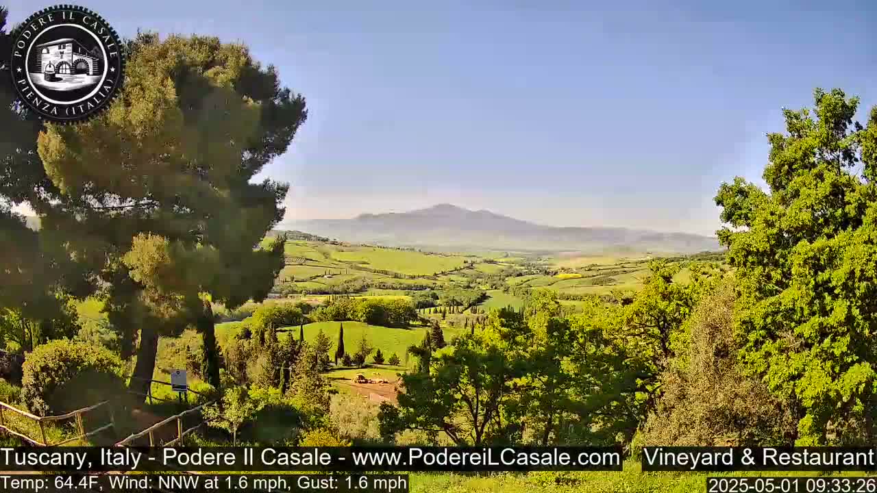 A picturesque, sunny Tuscan landscape unfolds, featuring rolling green hills, lush trees, and a distant mountain range under a clear sky.