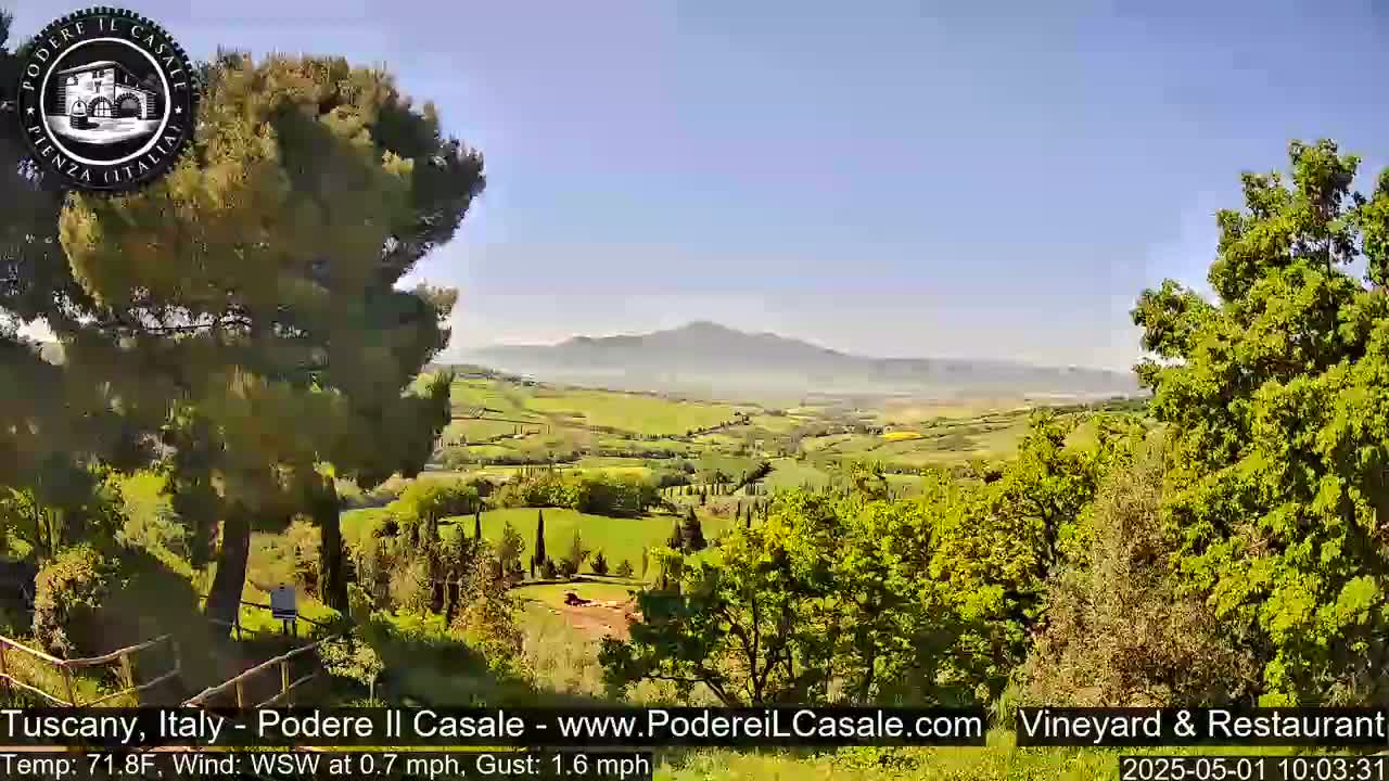 A panoramic view of a sun-drenched Tuscan landscape shows rolling green hills, trees, and a distant mountain range under a clear blue sky.