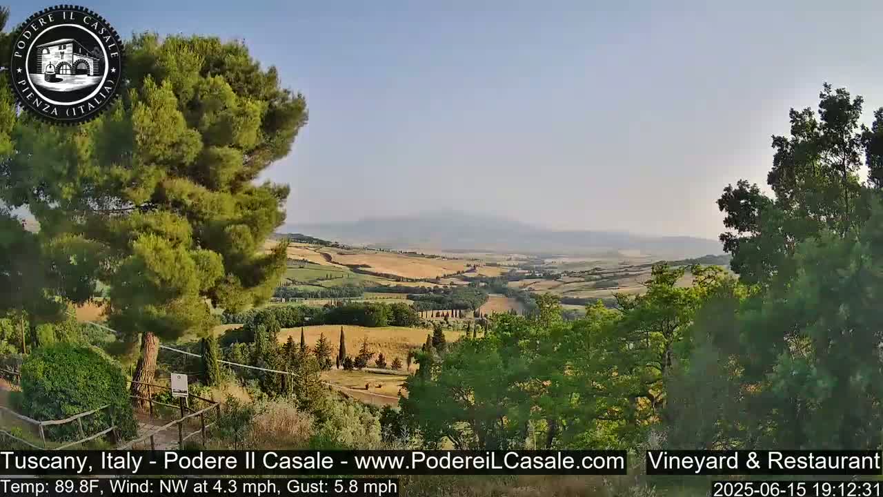 A panoramic view of a Tuscan landscape on a clear, warm day shows rolling hills of farmland, trees, and distant mountains under a light blue sky.