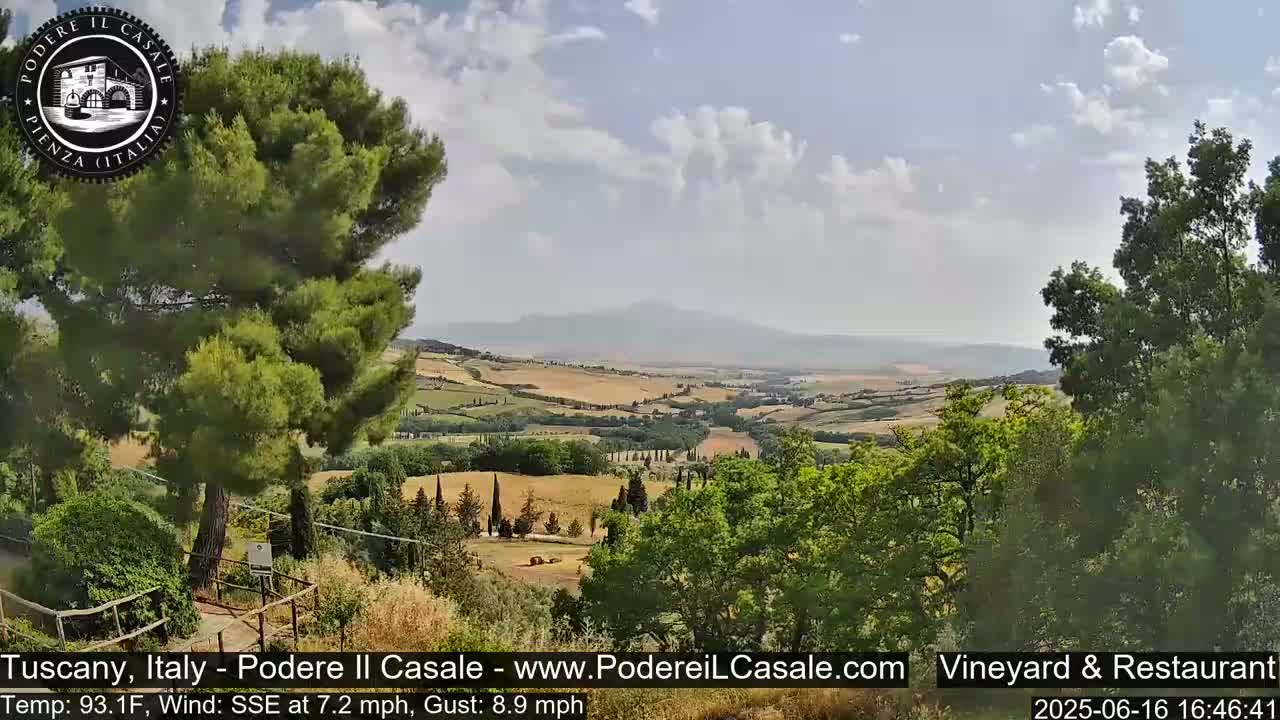 A partly cloudy Tuscan landscape, viewed from an elevated position, shows rolling hills dotted with trees and fields, under a light breeze.