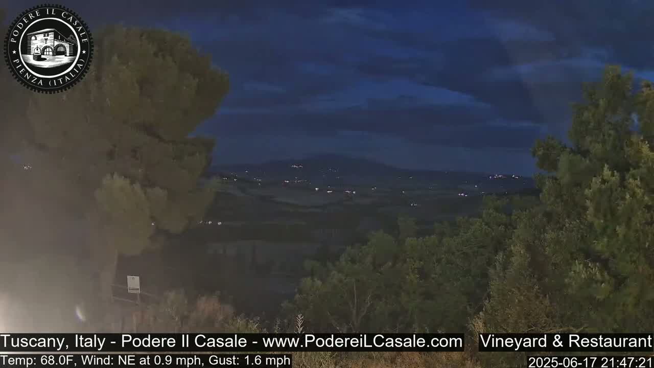 A nighttime view of a hilly landscape dotted with lights, under a dark blue, partly cloudy sky.