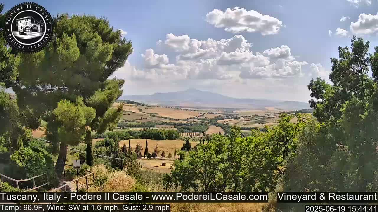 A partly cloudy sky overlooks a vast, rolling landscape of Tuscan hills and fields under a bright sun.