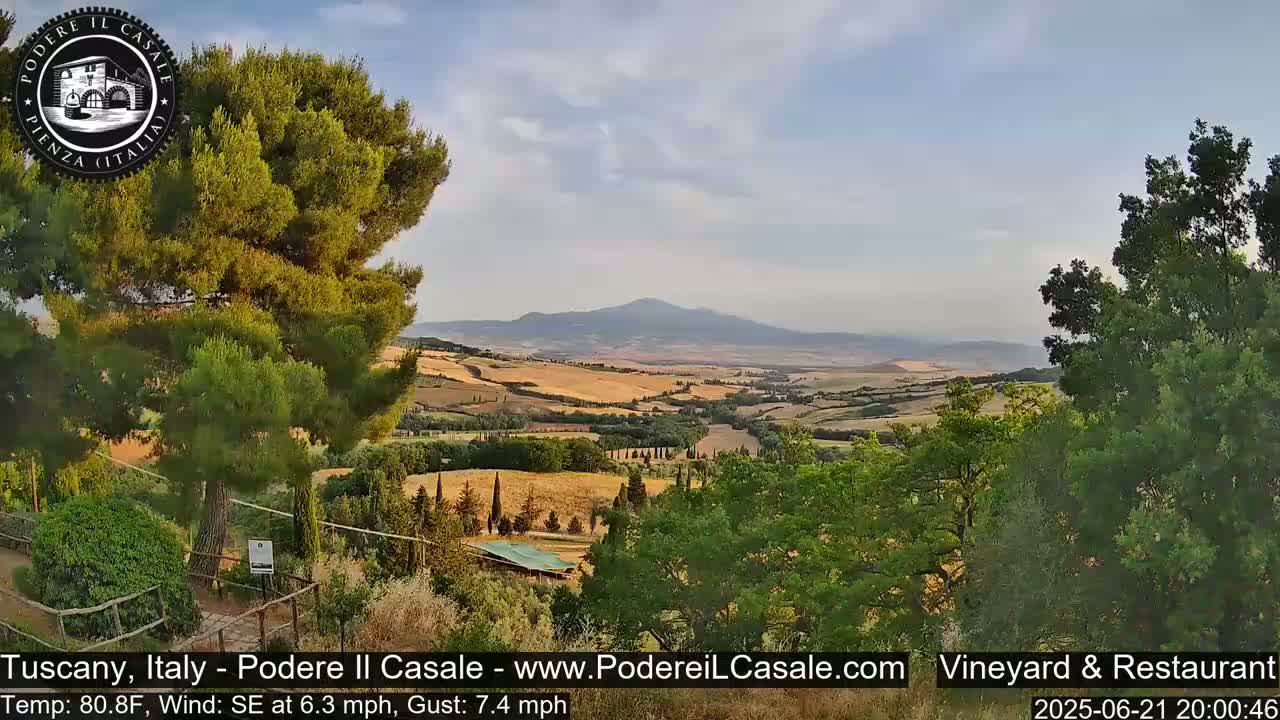 A picturesque Tuscan landscape under a partly cloudy sky shows rolling hills of golden-brown farmland, green trees, and a distant mountain range.