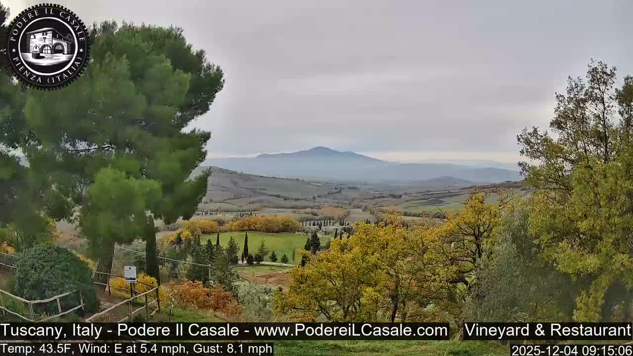 A wide view of a rolling rural landscape with fields and trees displaying autumn colors stretches towards distant hazy mountains under an overcast sky.