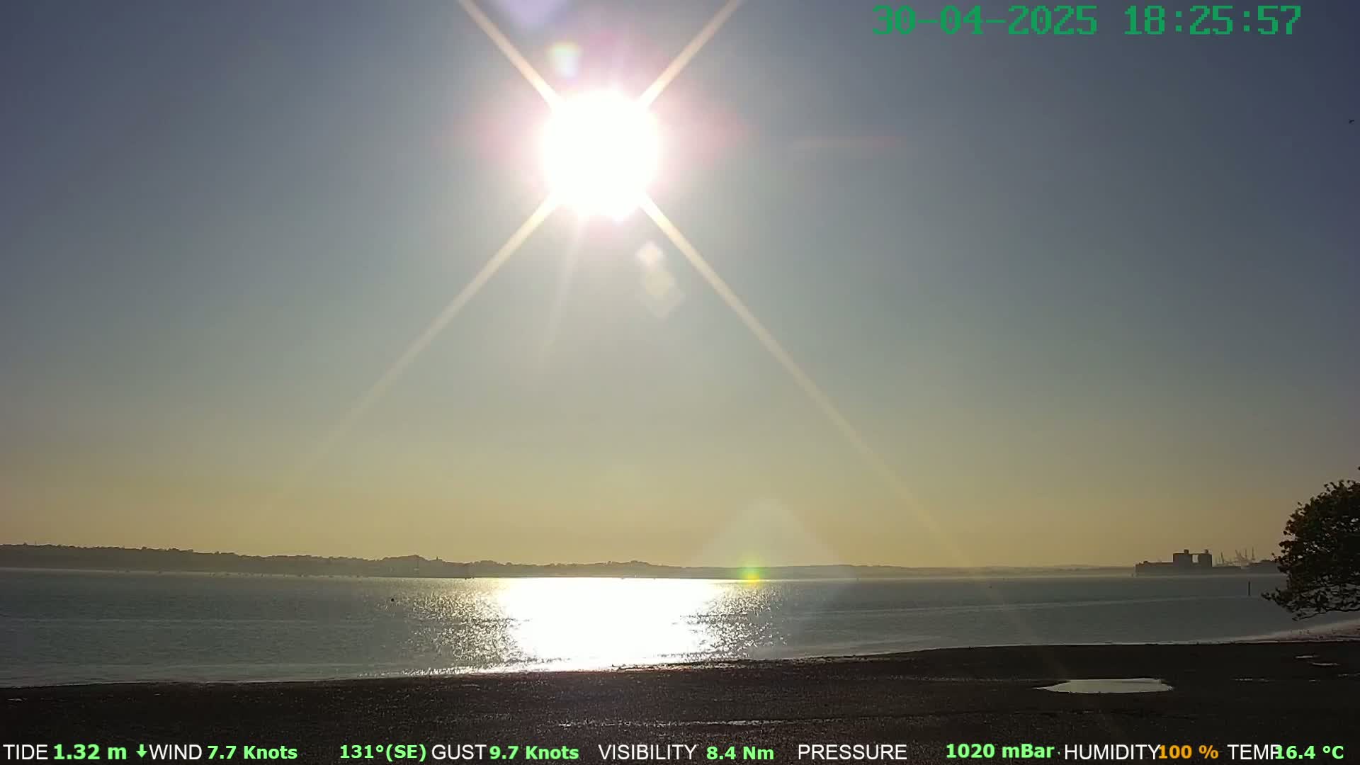 A bright sun shines over a calm body of water with a distant shoreline, under a mostly clear sky.