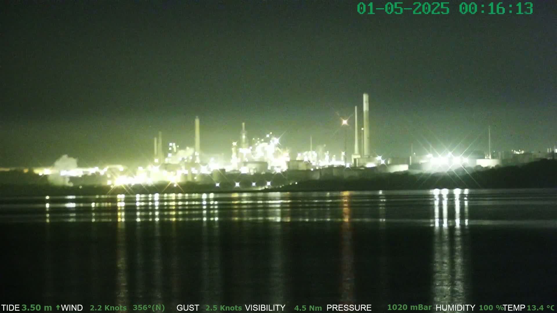 At night, a brightly lit industrial plant is reflected in calm water under a dark sky.