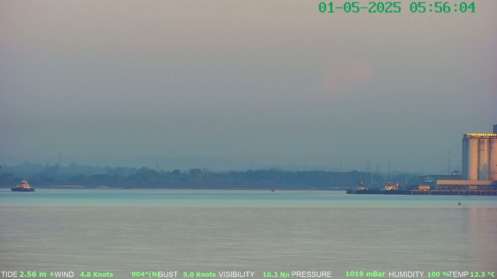A hazy, calm morning shows two tugboats on a body of water with a distant shoreline and industrial buildings.