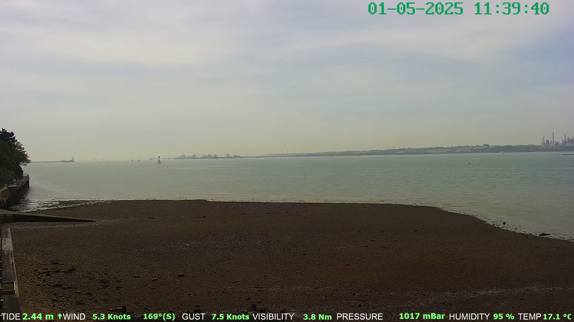 A hazy, overcast day reveals a calm body of water with a dark-colored, sandy beach in the foreground and an industrial shoreline in the distance.
