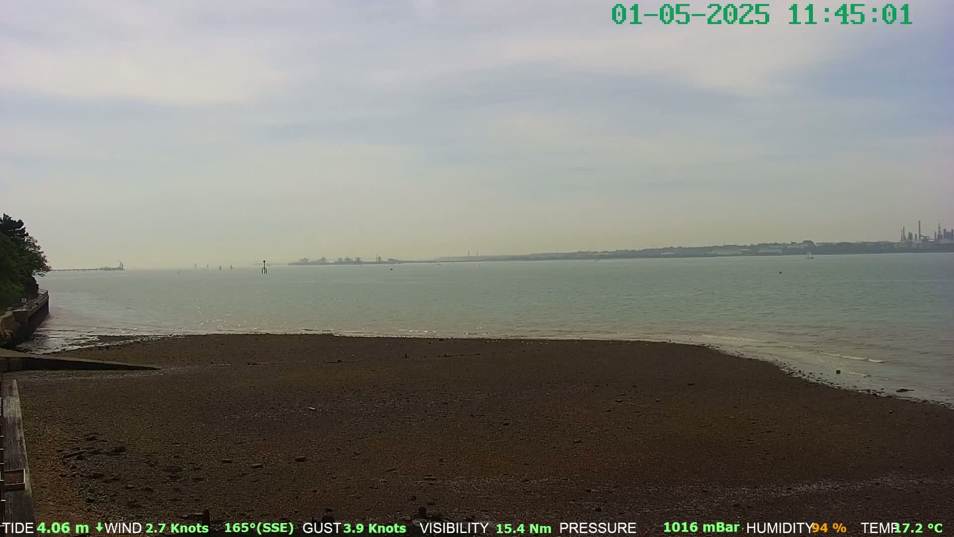 A mostly cloudy day reveals a wide, dark-brown sandy beach sloping gently to a calm, light-grey body of water, with a distant industrial shoreline visible on the horizon.
