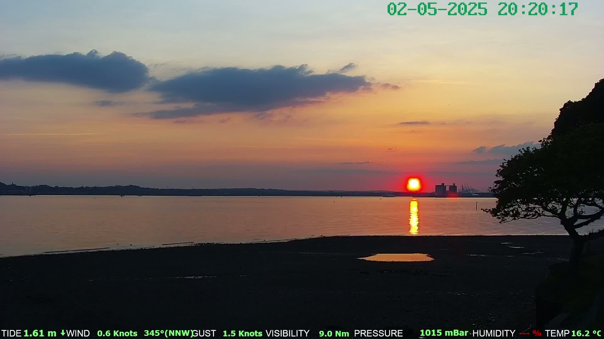 A large, orange sun sets over calm water, casting a reflection on a dark, sandy shore beneath a partly cloudy sky.