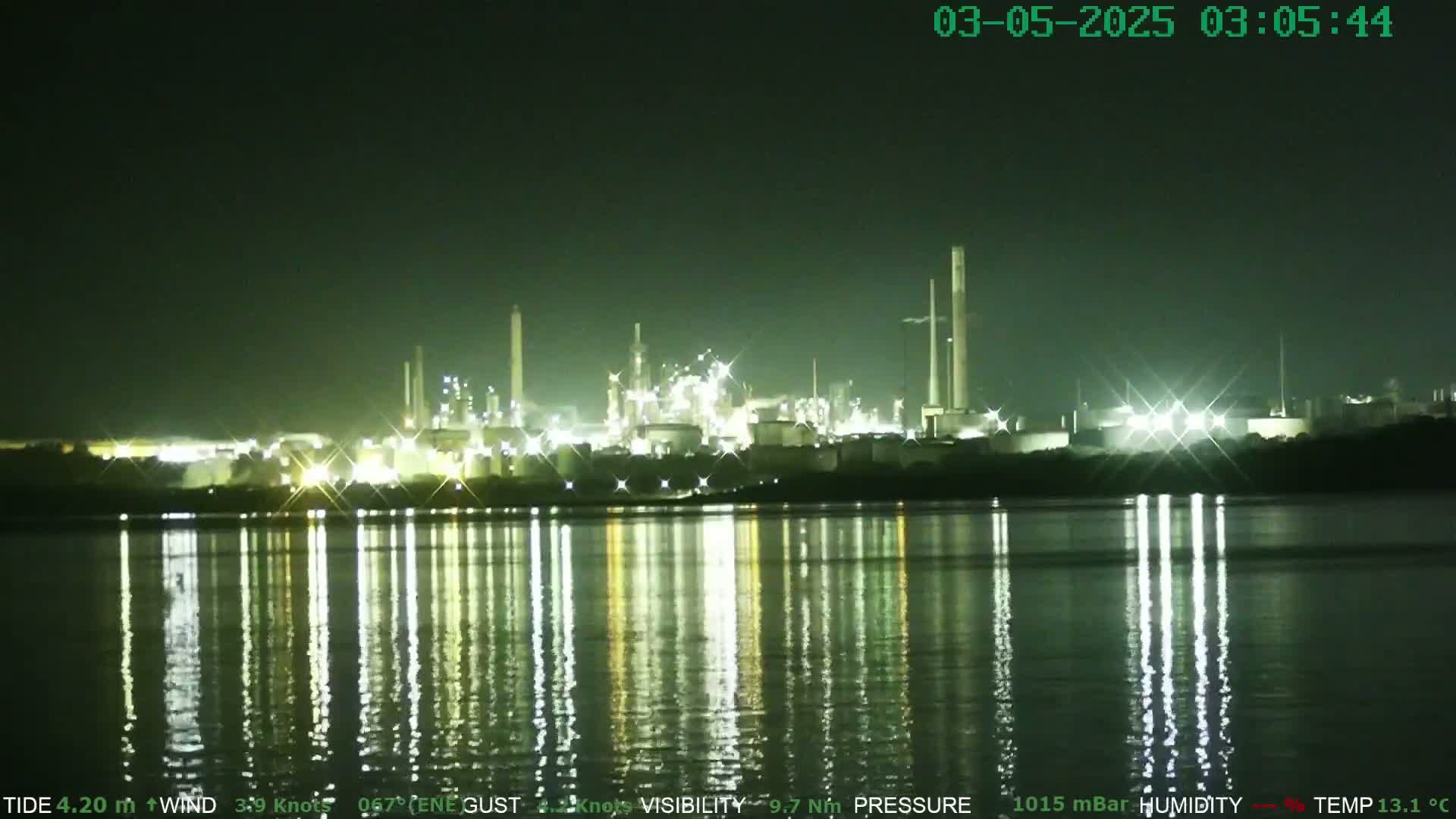 A brightly lit industrial complex is reflected in the calm waters of a body of water at night.