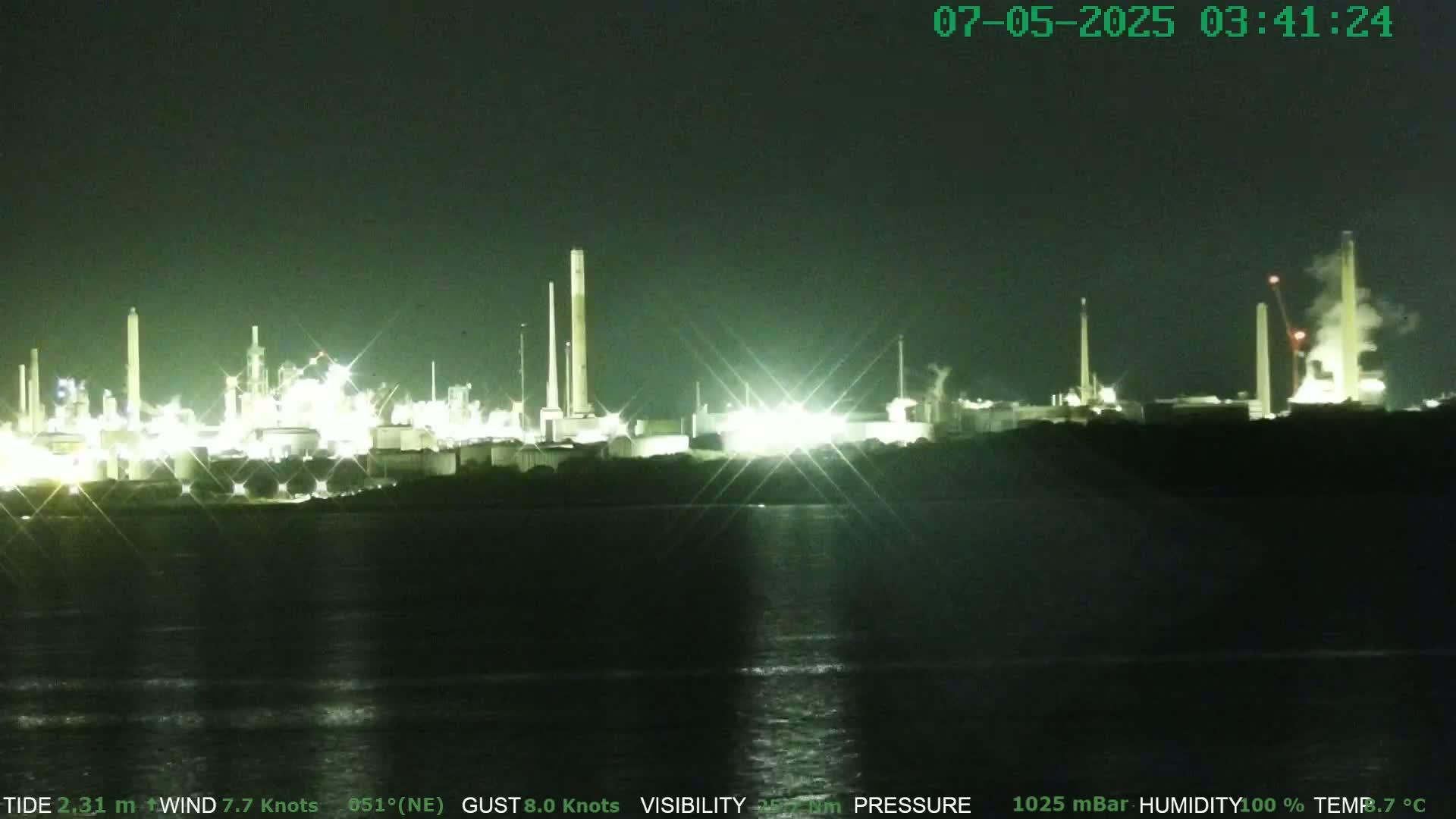 A brightly lit industrial plant at night is seen across a dark body of water under a clear, dark sky.