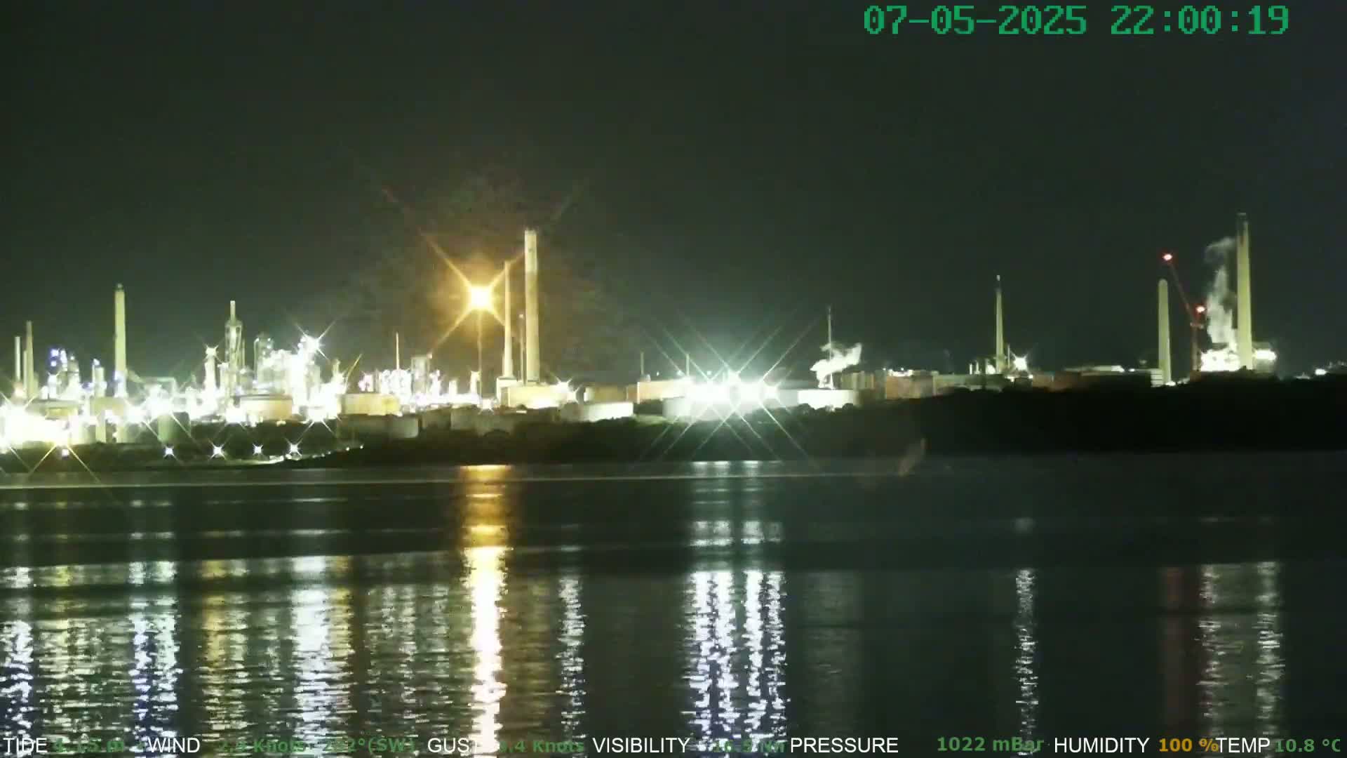 A brightly lit industrial complex is situated across a calm body of water at night.