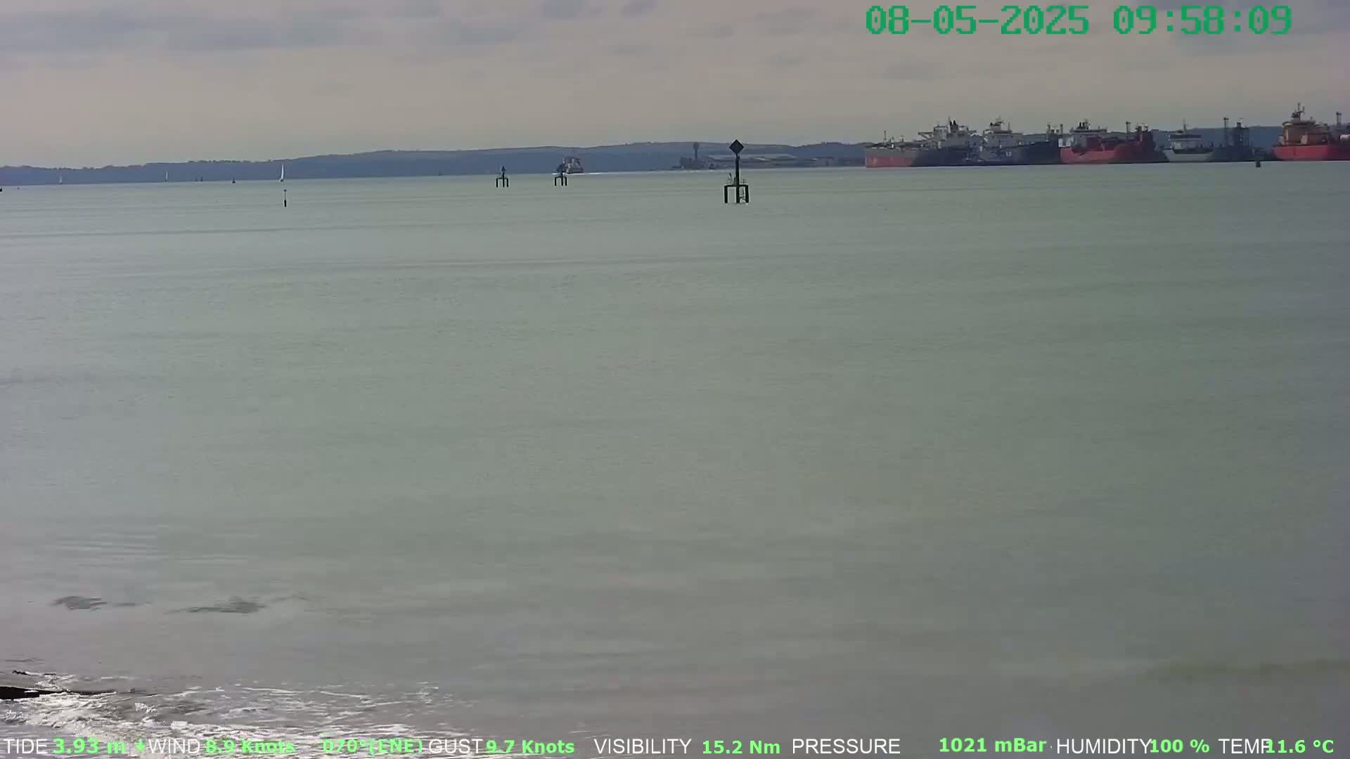 A calm, overcast day shows several ships in a harbor, with a few sailboats and navigation markers visible on the water.