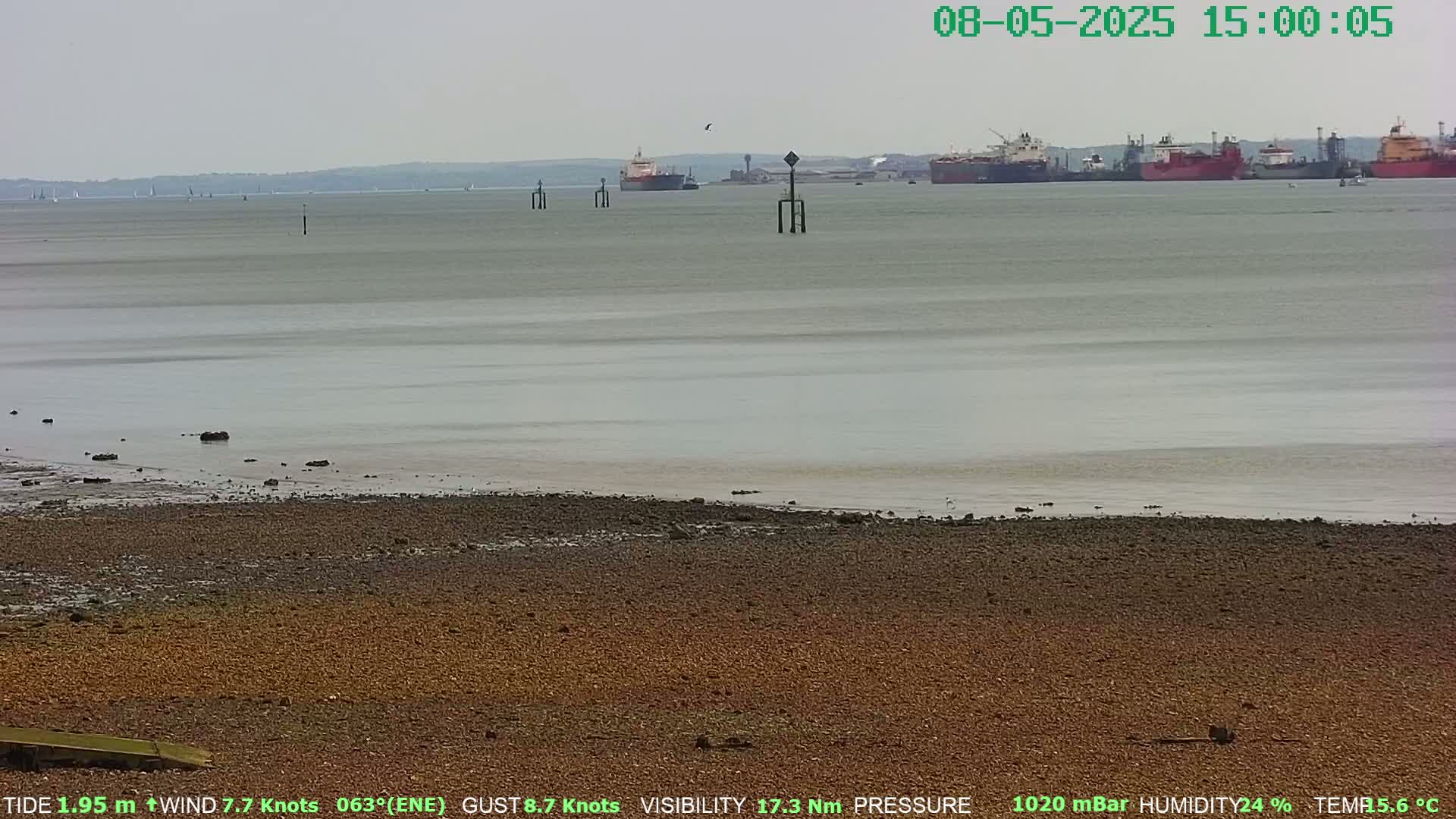 A wide shot of a calm, grey-watered body of water with several cargo ships in the distance, a pebbled shoreline in the foreground under an overcast sky.