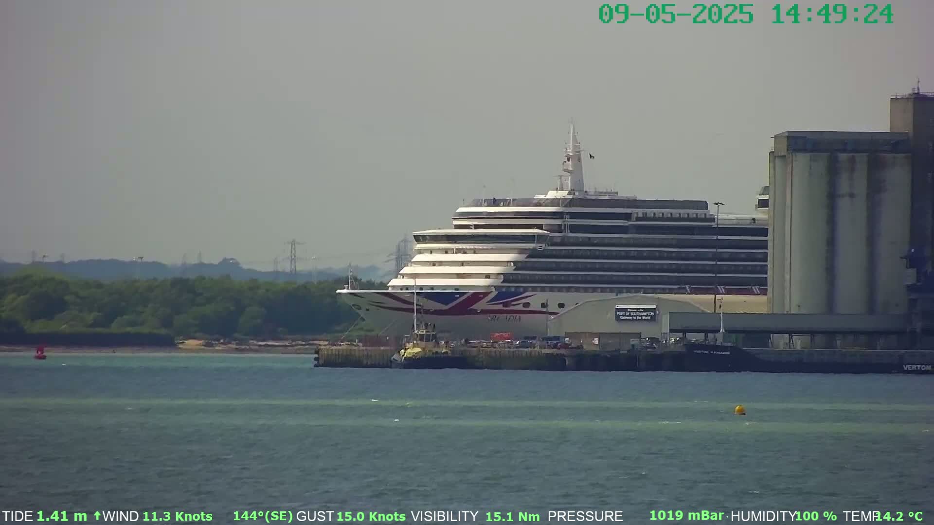 A large cruise ship is docked near industrial buildings on a hazy day with a 11.3 knot wind and 15.1 nm visibility.