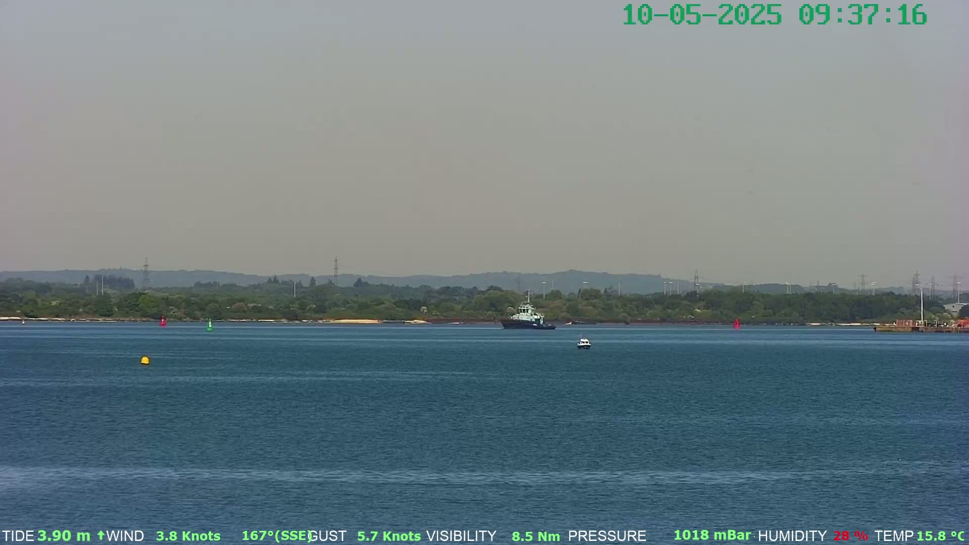A tugboat and a smaller vessel are on a calm, hazy body of water in front of a distant shoreline of green trees and some structures.