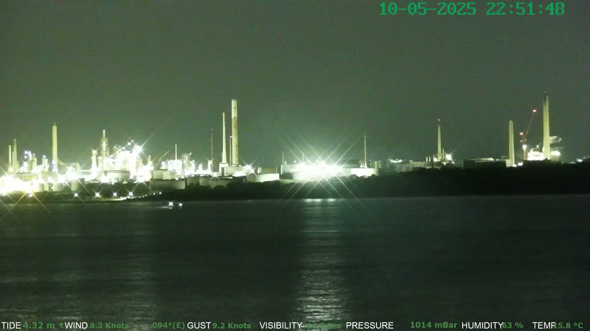A brightly lit industrial plant sits on a dark shoreline across a calm body of water at night.