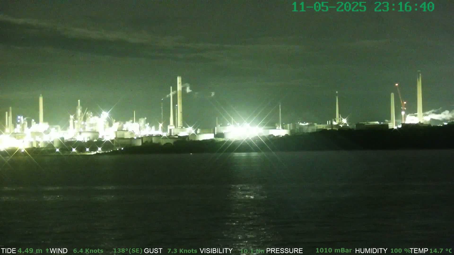 A brightly lit industrial complex is visible across a dark body of water at night under a mostly clear sky.