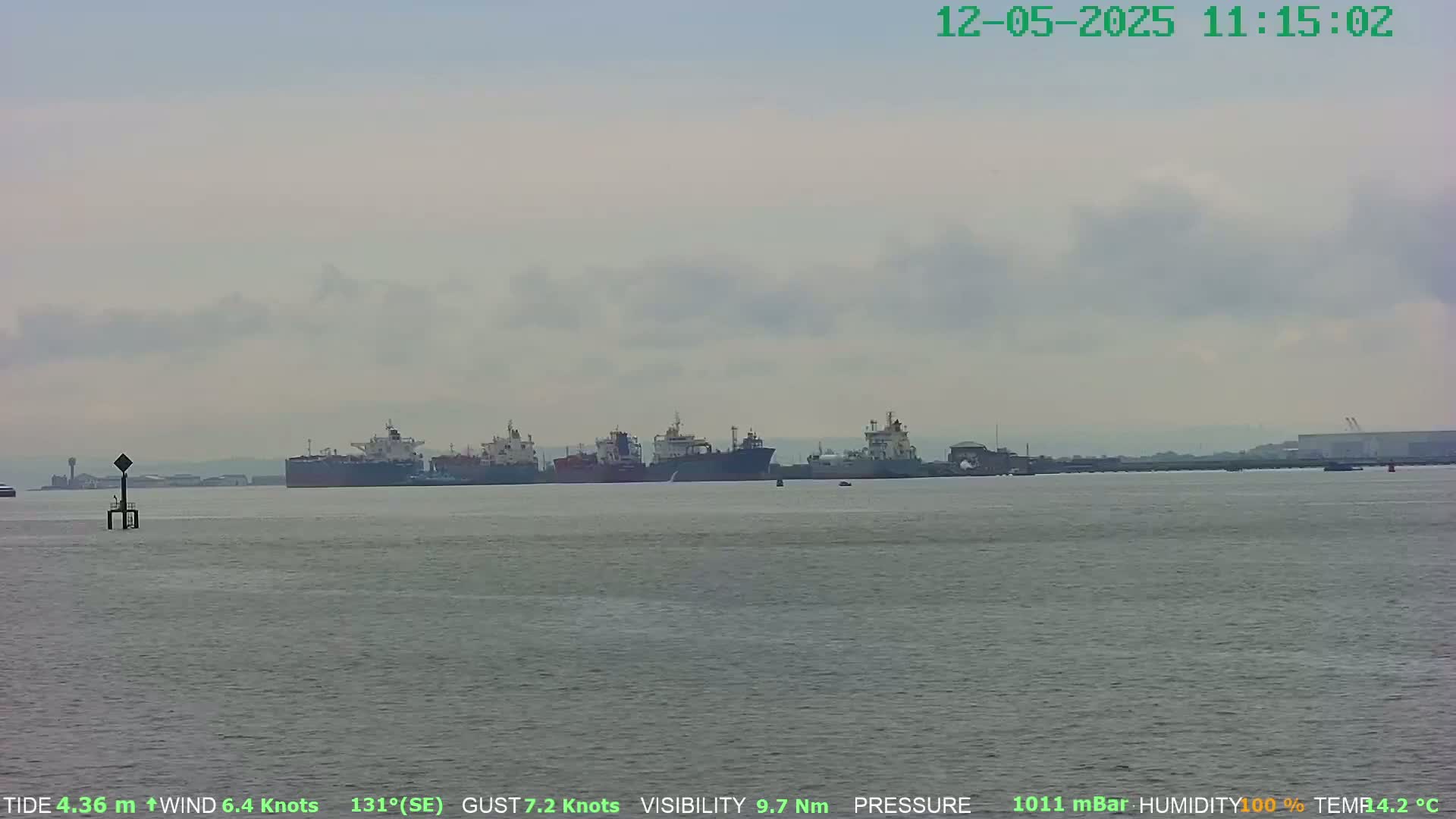 A calm, overcast day shows several large ships at anchor in a body of water, with a navigational marker in the foreground.