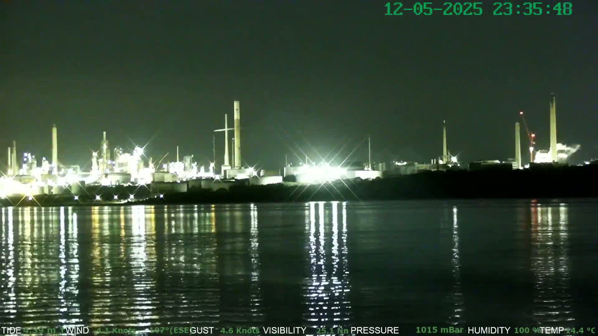 At night, a brightly lit industrial complex sits on the far shore of a calm, dark body of water, its lights reflecting on the surface.