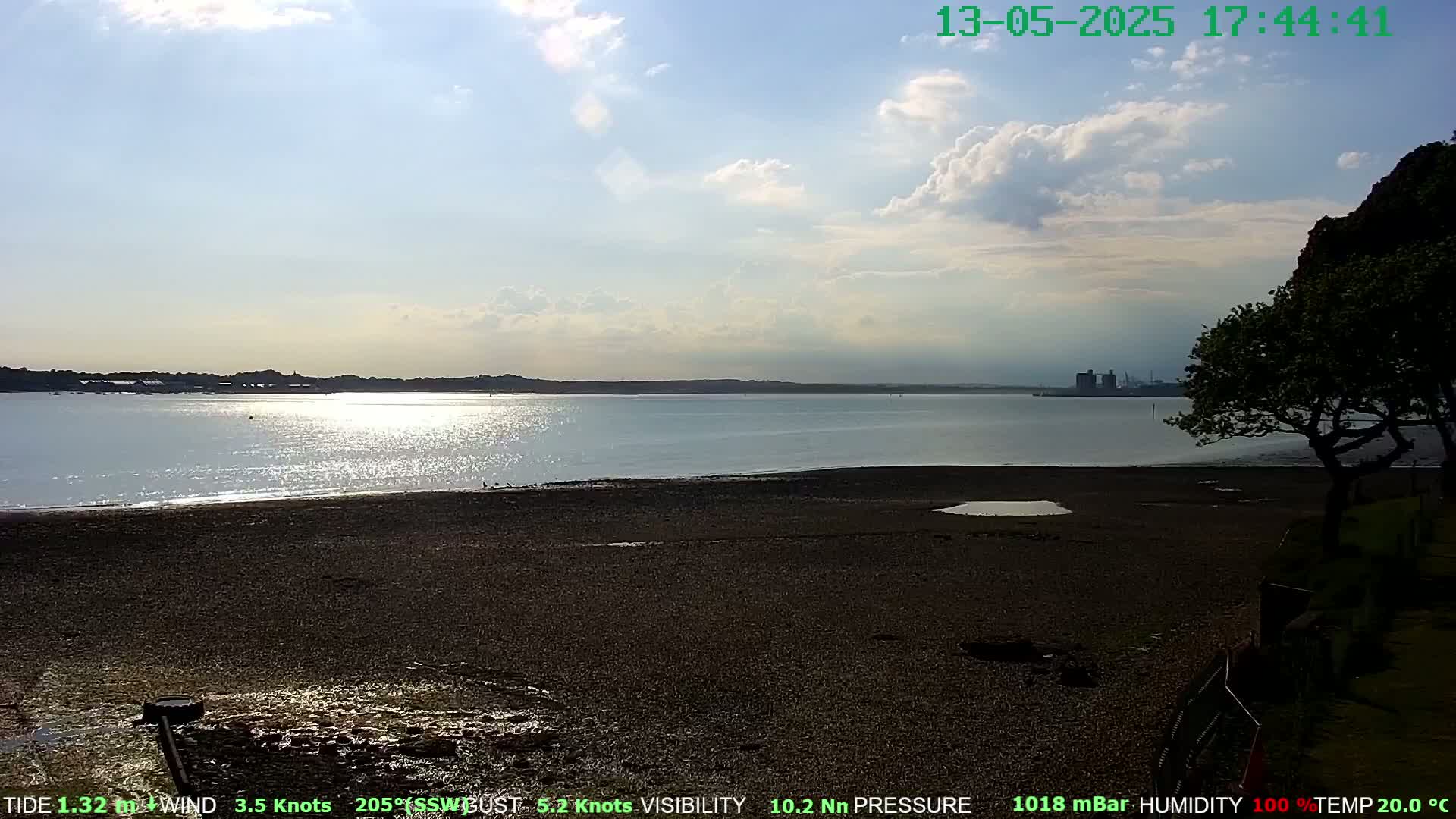 A partly cloudy afternoon scene shows a calm body of water reflecting sunlight, a dark-colored sandy beach, and a tree silhouetted against the background.