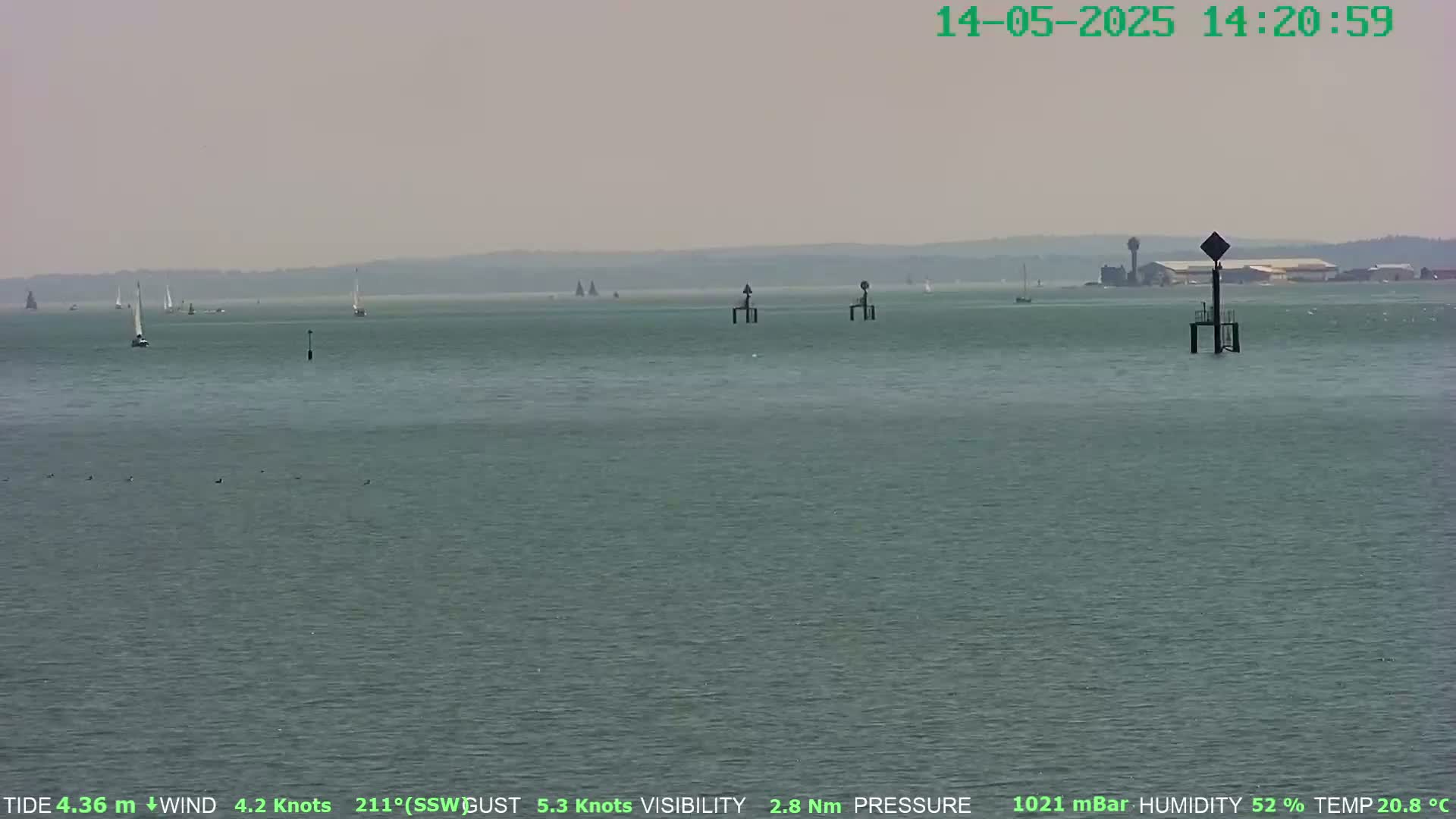 A hazy, calm sea with several sailboats in the distance and navigational markers in the foreground, under a light grey sky.