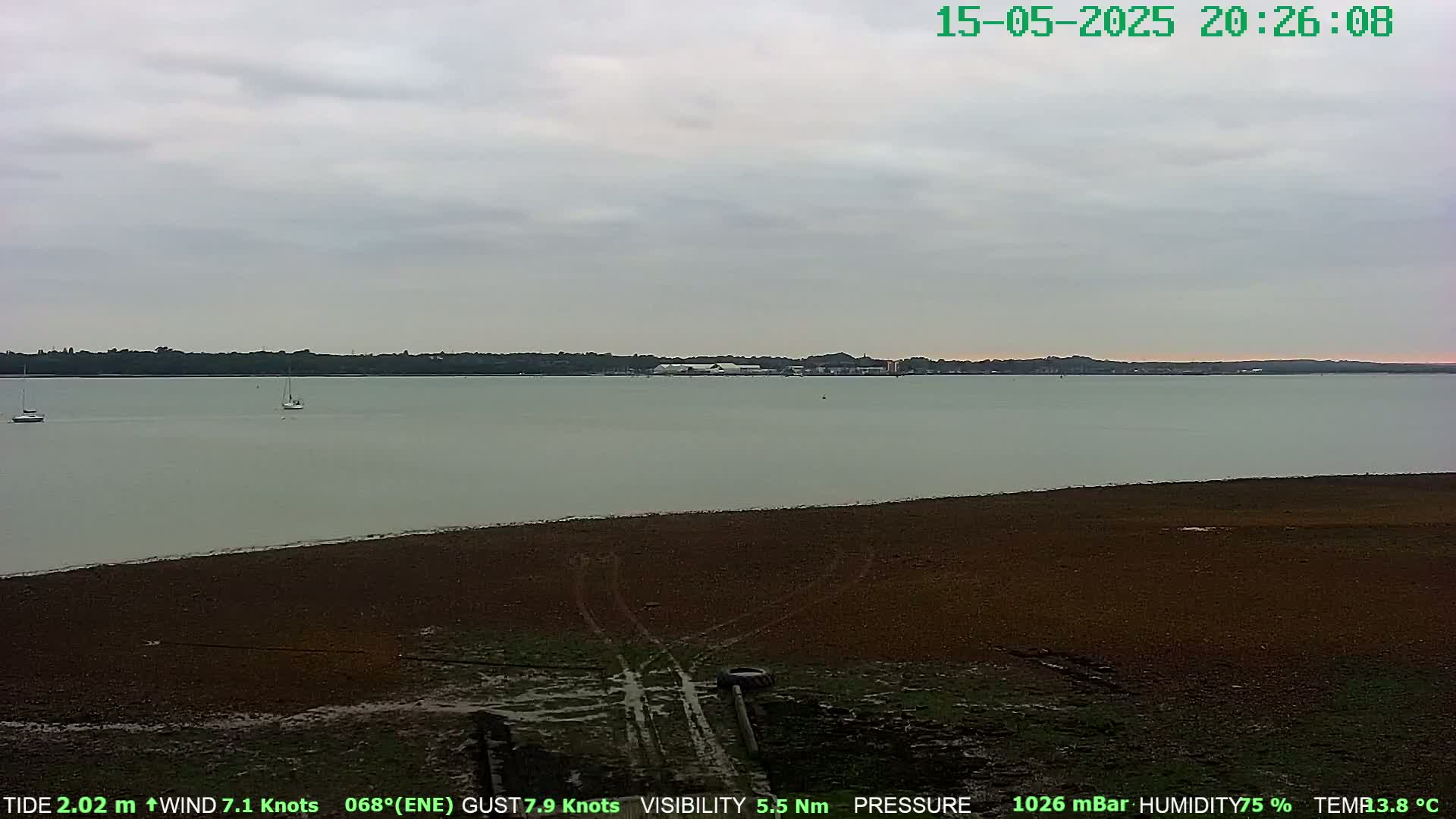 A calm, overcast body of water is seen from a muddy shoreline with tire tracks, and two small sailboats are visible in the distance.