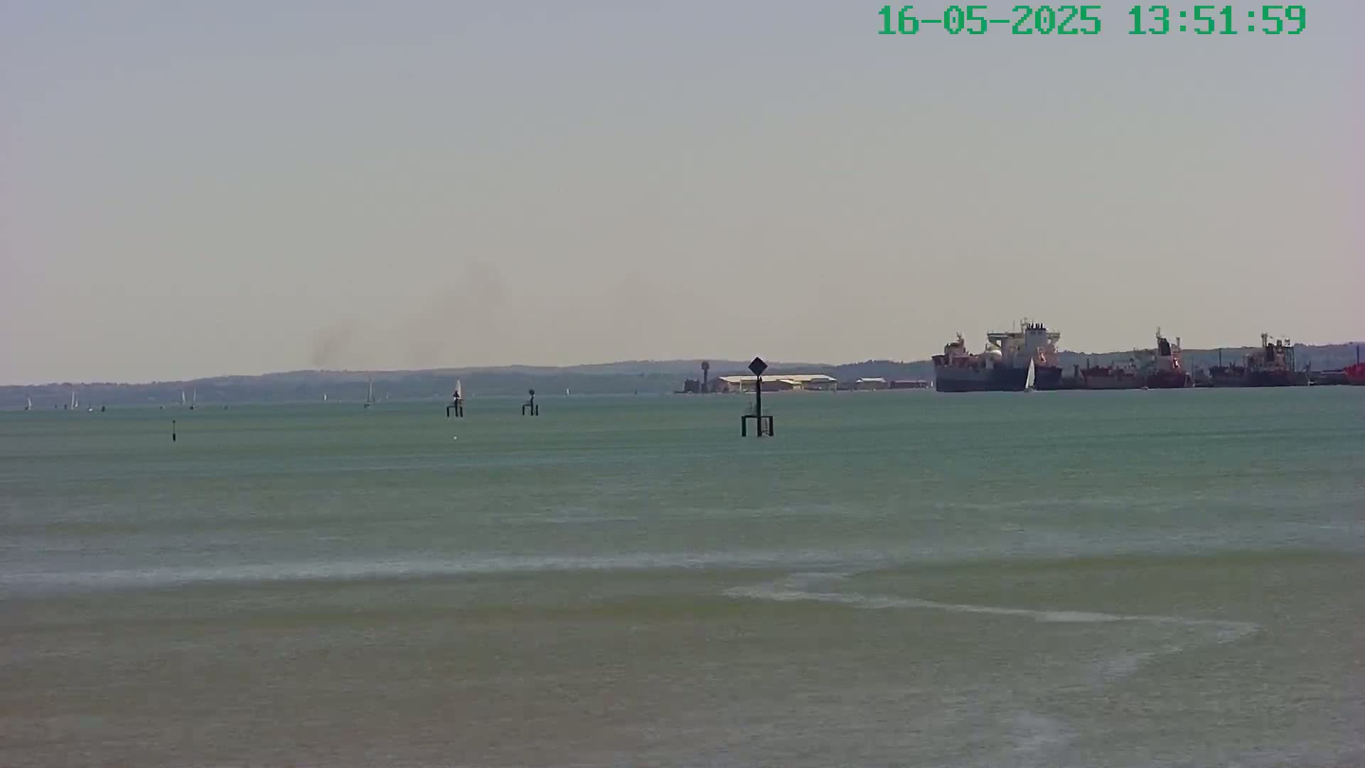 Several ships are visible on a calm, hazy day at sea, with a few sailboats and navigational markers in the distance.