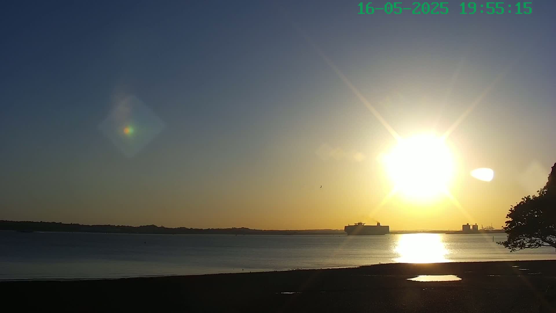 A bright sun sets over calm water, silhouetting a large ship and distant shoreline under a clear sky.