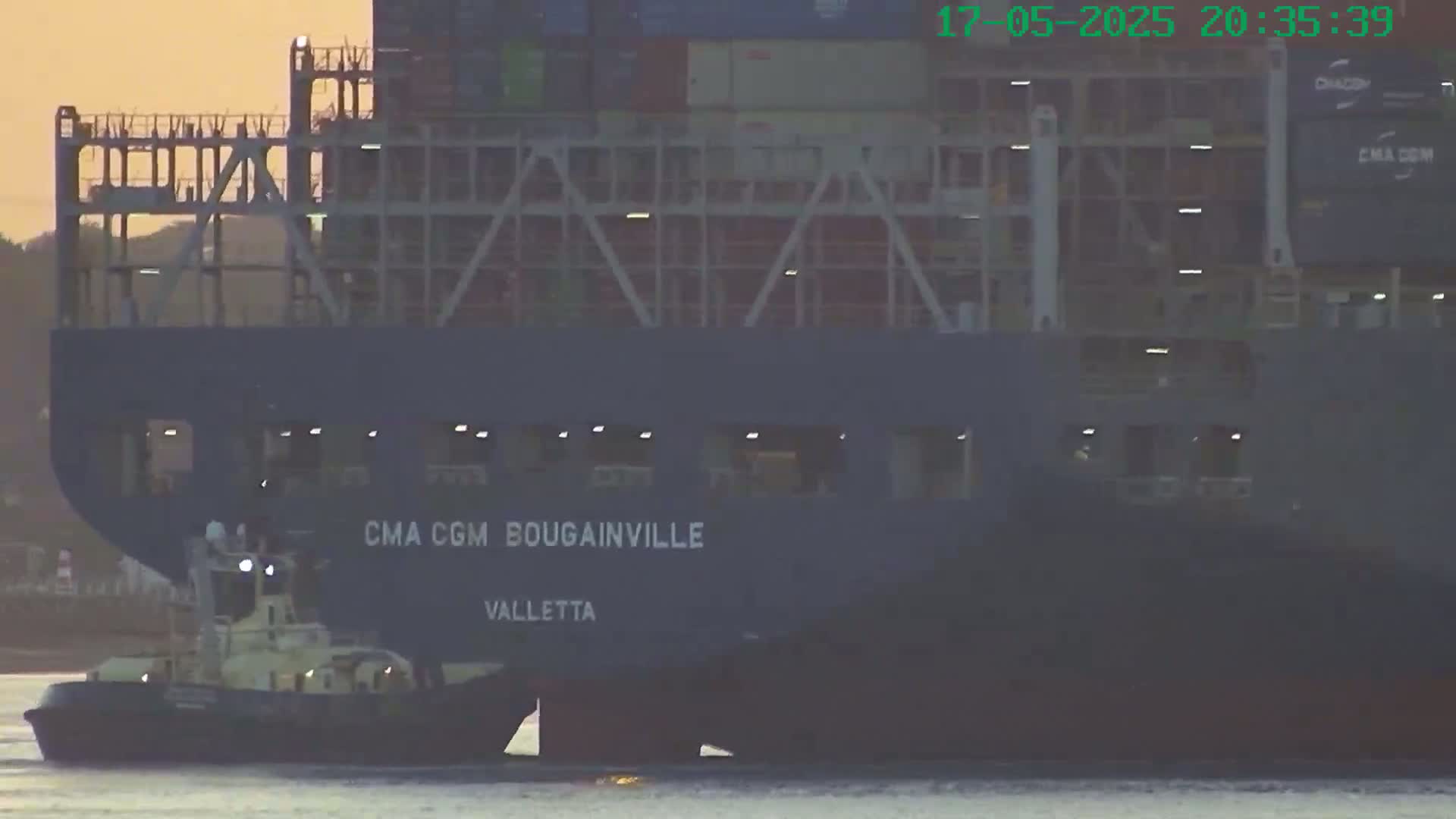A large container ship named CMA CGM Bougainville, registered in Valletta, is being assisted by a tugboat in calm, dusky-orange light.
