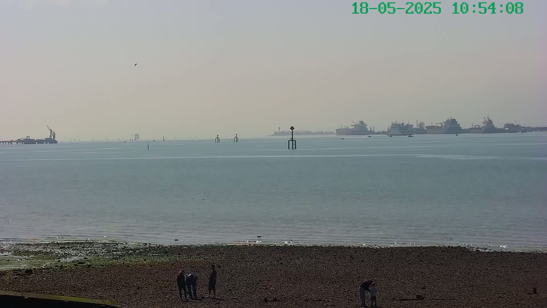 A hazy, sunny day reveals a calm body of water with a pebbled beach in the foreground, where several people stand, and industrial structures and ships in the distance.