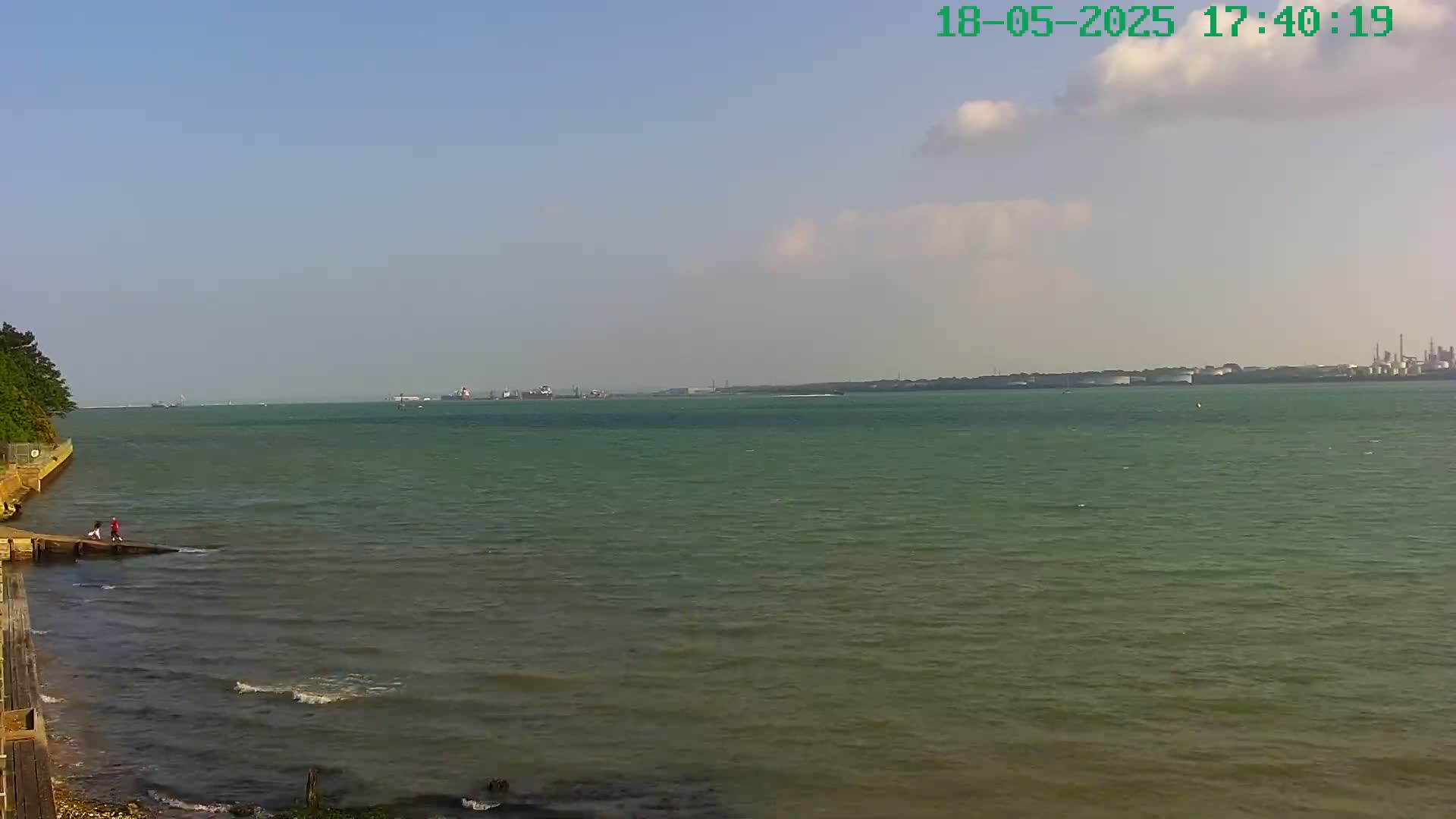 A calm, partly cloudy day shows a wide body of water with a shoreline featuring a small group of people near a short pier, distant ships, and an industrial area visible on the horizon.