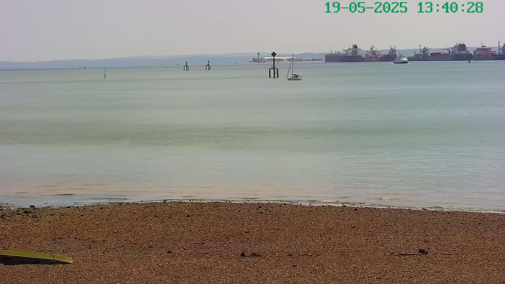A calm, light-grey body of water with several ships in the distance and a sailboat closer to shore, viewed from a sandy beach under a hazy sky.