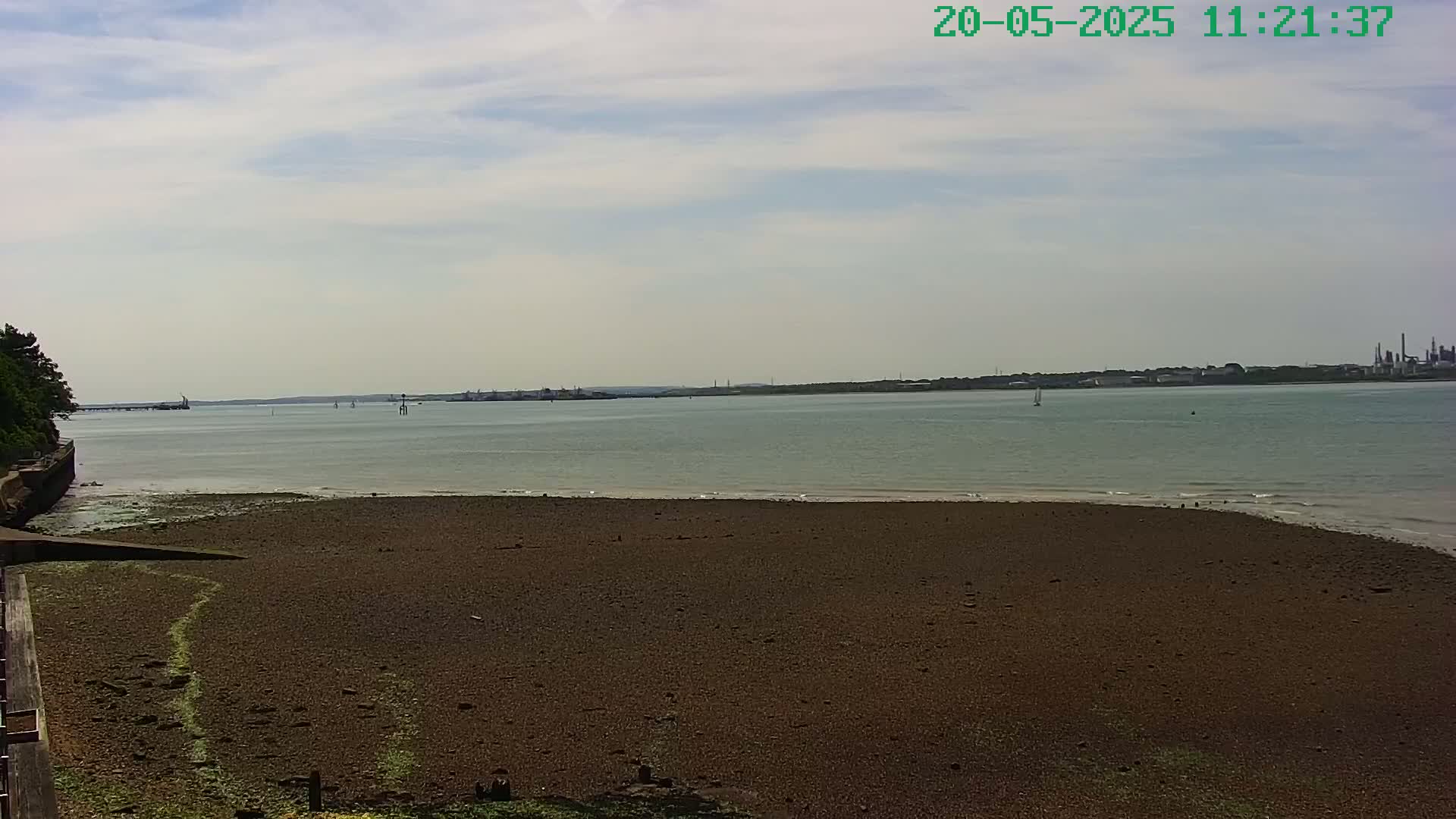 A mostly sunny day shows a wide expanse of dark-brown mudflats stretching to a calm body of water with a distant shoreline under a partly cloudy sky.