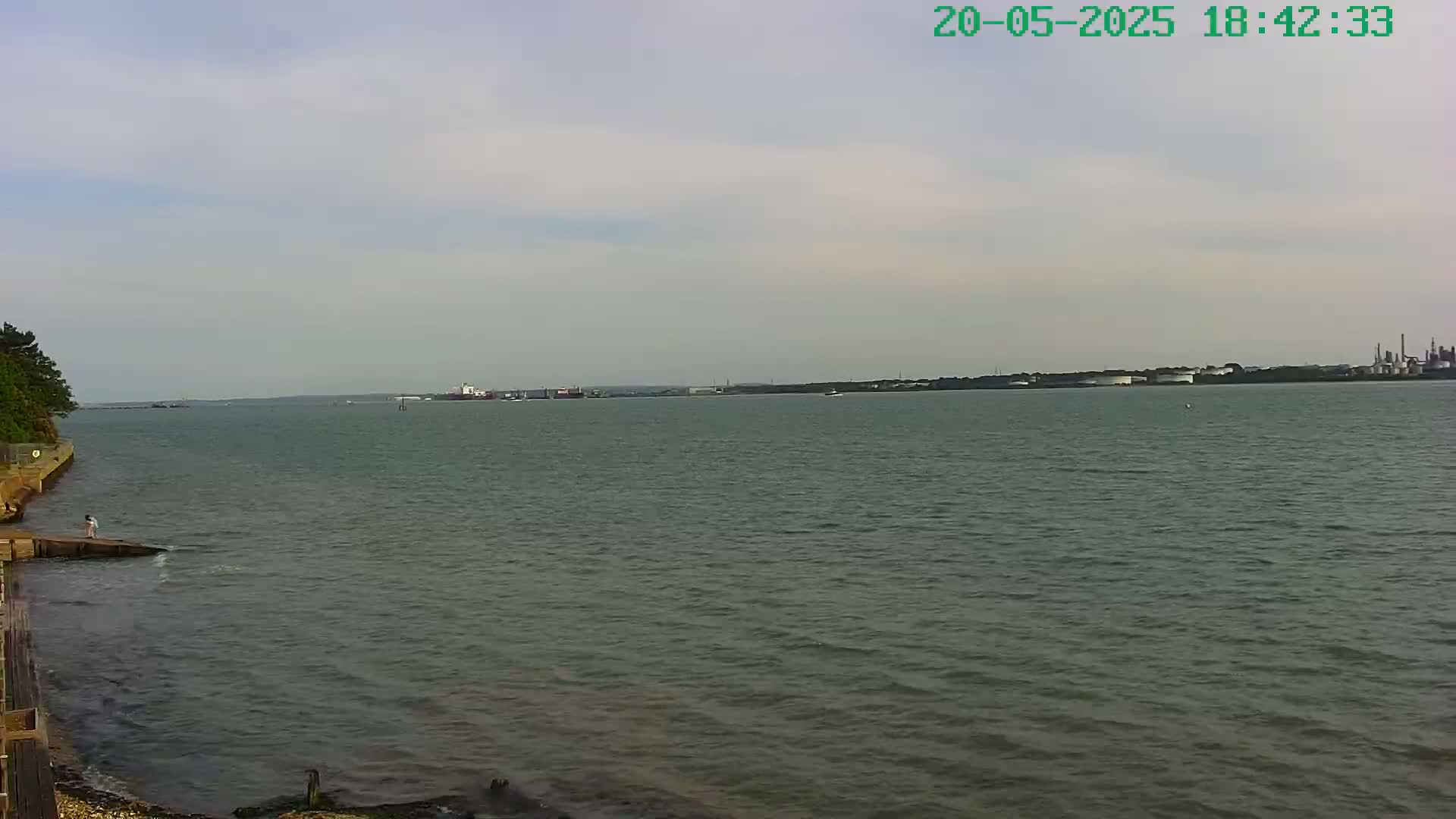 A calm, overcast day reveals a wide body of water with a distant industrial shoreline and a small figure near a wooden structure on the left.