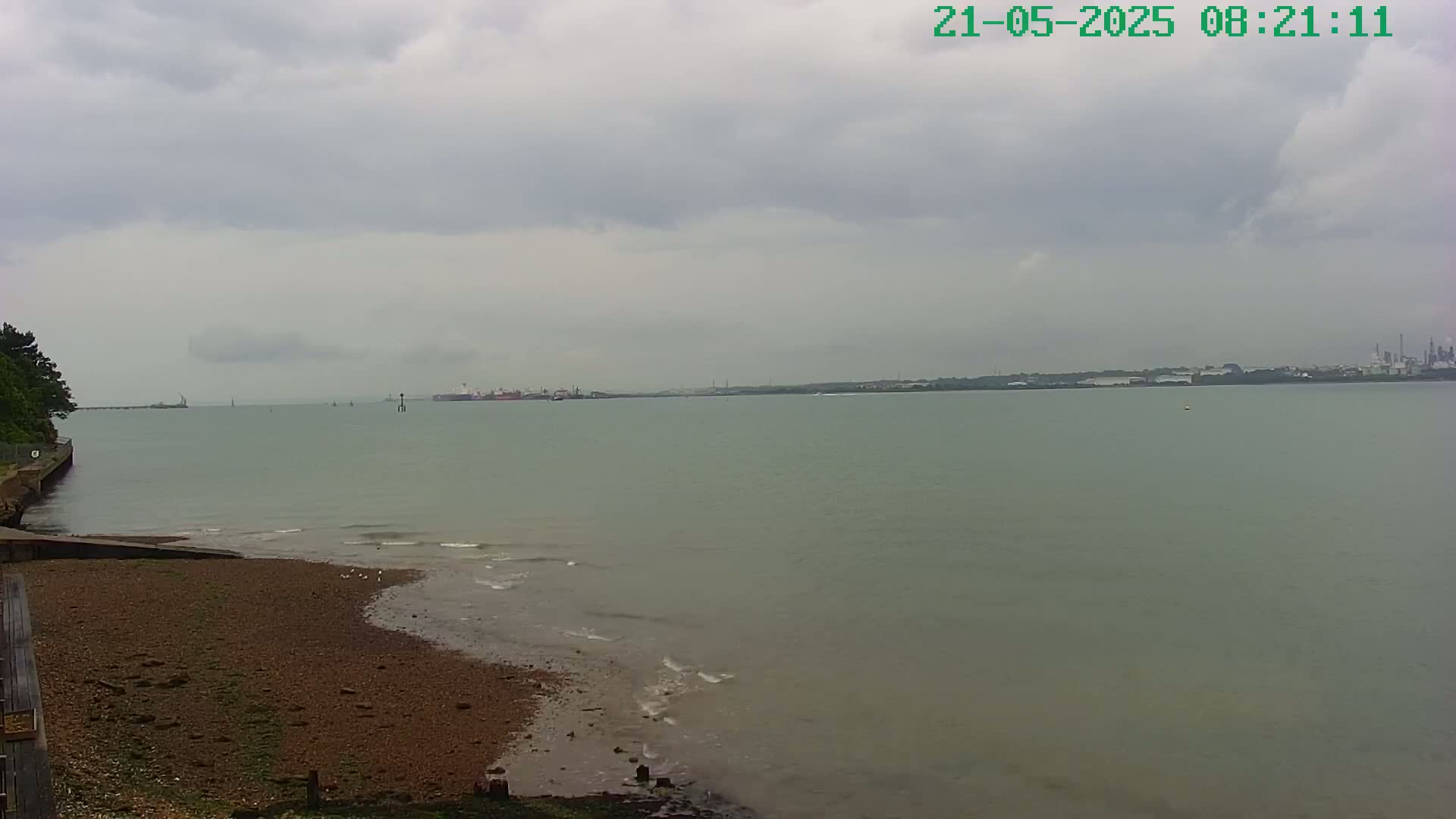 A calm, overcast day shows a pebbled shoreline leading to a body of water with distant industrial structures visible on the horizon.